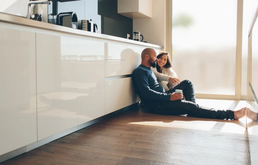 A couple sitting on the floor of a modern white kitchen leaning against the cabinets talking and smiling